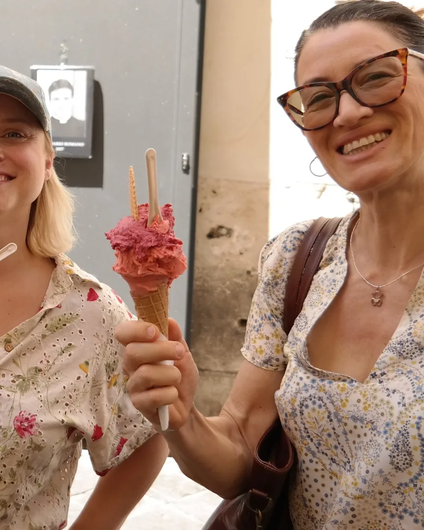 Two guests smiling on a Roman street, one holding up a cone of raspberry gelato with a wafer in it.