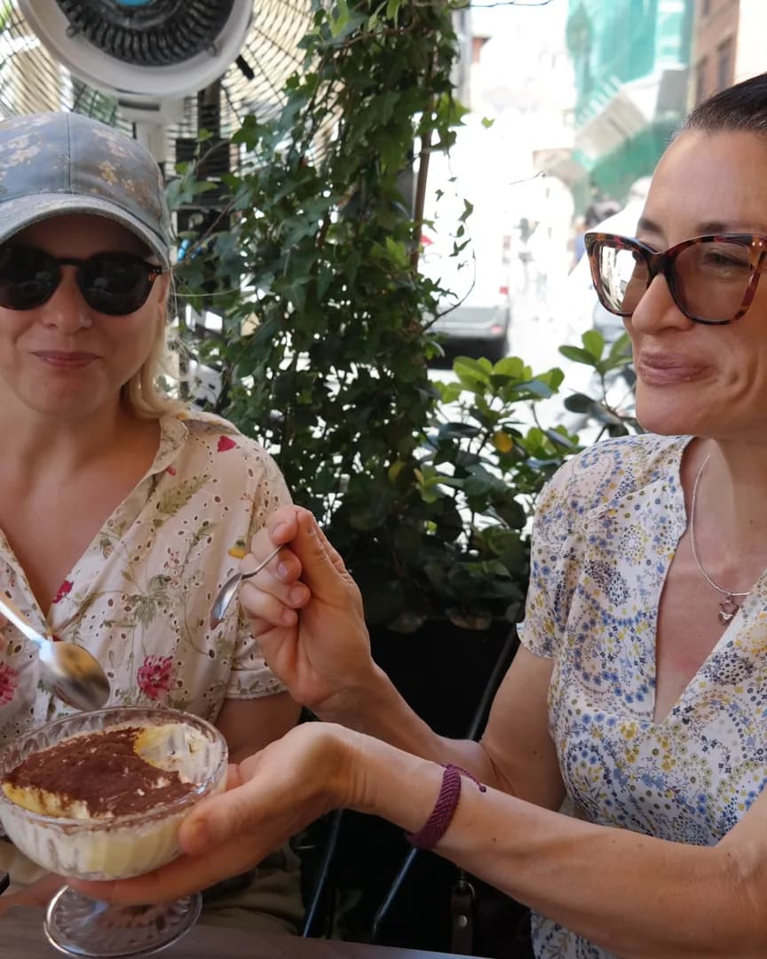 Two guests at a shaded pavement table sharing a glass bowl of tiramisu, spoons in hand.